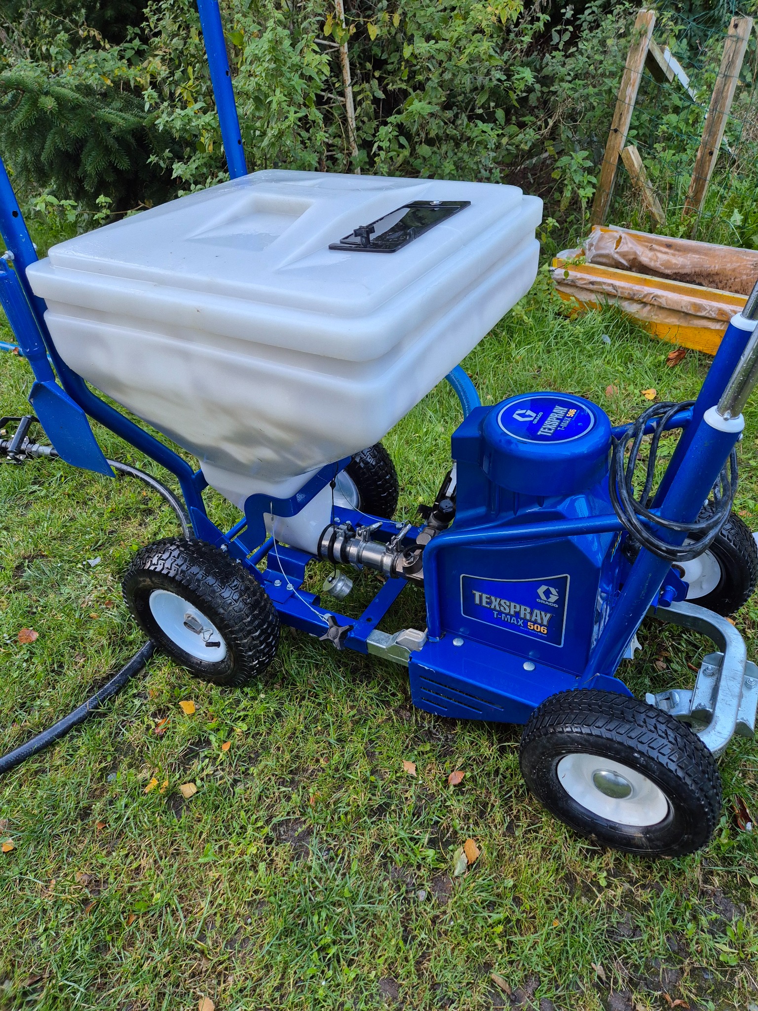 Agregat malarski TexSpray T-Max 506 na trawie, z białym zbiornikiem i niebieską konstrukcją. Widoczne koła i przewód. Sprzęt gotowy do użycia na zewnątrz.