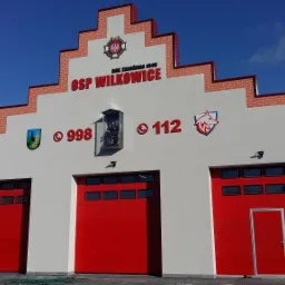 Fasada budynku Ochotniczej Straży Pożarnej w Wilkowicach z trzema czerwonymi bramami garażowymi, oznaczona numerami alarmowymi 998 i 112, herbem i nazwą OSP Wilkowice na tle błękitnego nieba.