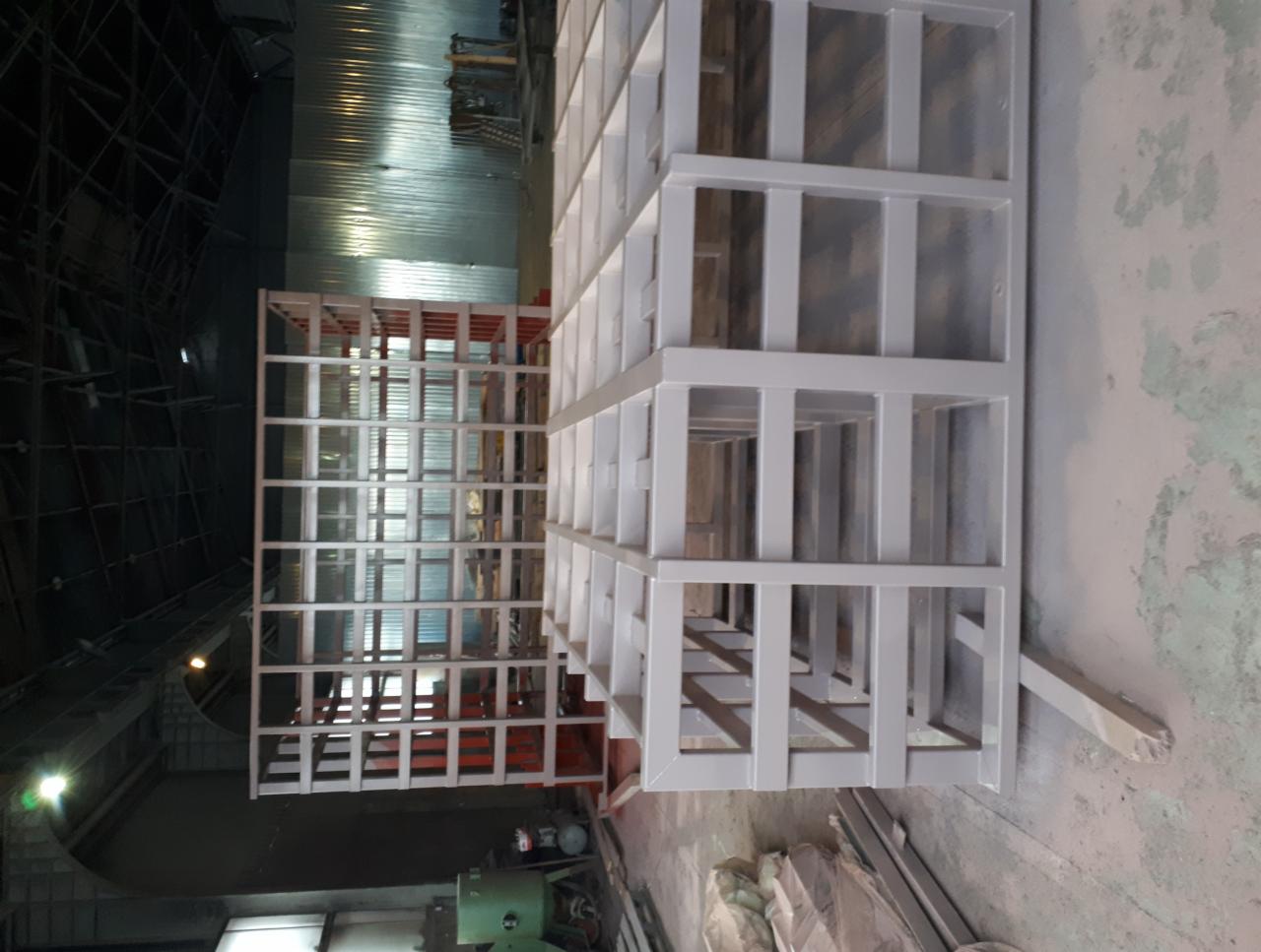 Metalowe konstrukcje kratowe, pomalowane proszkowo na szaro, ustawione w hali produkcyjnej z widocznym oświetleniem sufitowym i elementami maszyn.