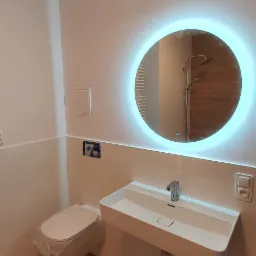 Nowoczesna łazienka z drewnopodobną podłogą, podwieszana umywalka z baterią i okrągłe lustro z podświetleniem LED; toaleta w folii ochronnej.