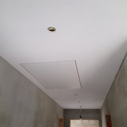 K.B. BUD - Szare, surowe ściany korytarza w trakcie remontu, kontrastujące z gładkim, białym sufitem z widocznym włazem rewizyjnym i otworem na oświetlenie.