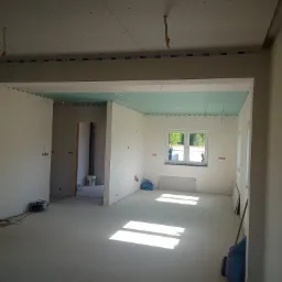 Wnętrze w trakcie remontu: ściany przygotowane do malowania z widocznymi punktami elektrycznymi, sufit częściowo obłożony płytami, podłoga z wylewką samopoziomującą, widoczne narzędzia i materiały...