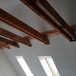K.B. BUD - Poddasze po remoncie z widocznymi drewnianymi belkami stropowymi i dwoma oknami połaciowymi, ściany i sufit pomalowane na biało. W tle widoczna żarówka na kablu.