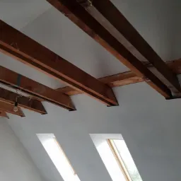 Poddasze po remoncie z widocznymi drewnianymi belkami stropowymi i dwoma oknami połaciowymi, ściany i sufit pomalowane na biało. W tle widoczna żarówka na kablu.