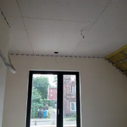 Wnętrze w trakcie remontu, widoczne zamontowane płyty na suficie, zabezpieczone taśmą, z widocznym otworem na oświetlenie, oraz fragment poddasza z izolacją, okno z widokiem na ulicę i budynki.