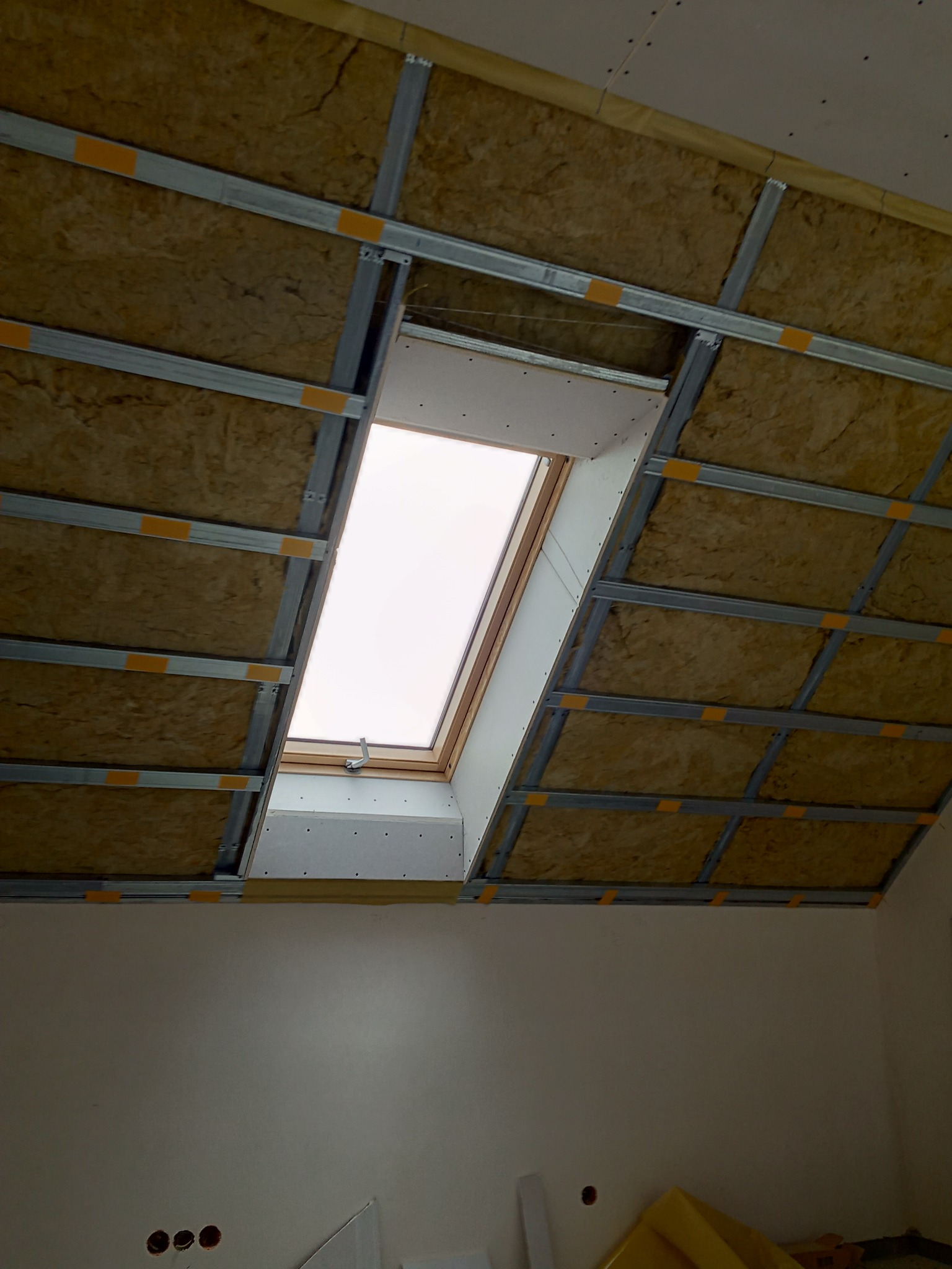 Montaż izolacji z wełny mineralnej i metalowego stelaża pod przyszłą zabudowę sufitu, widoczne okno dachowe.