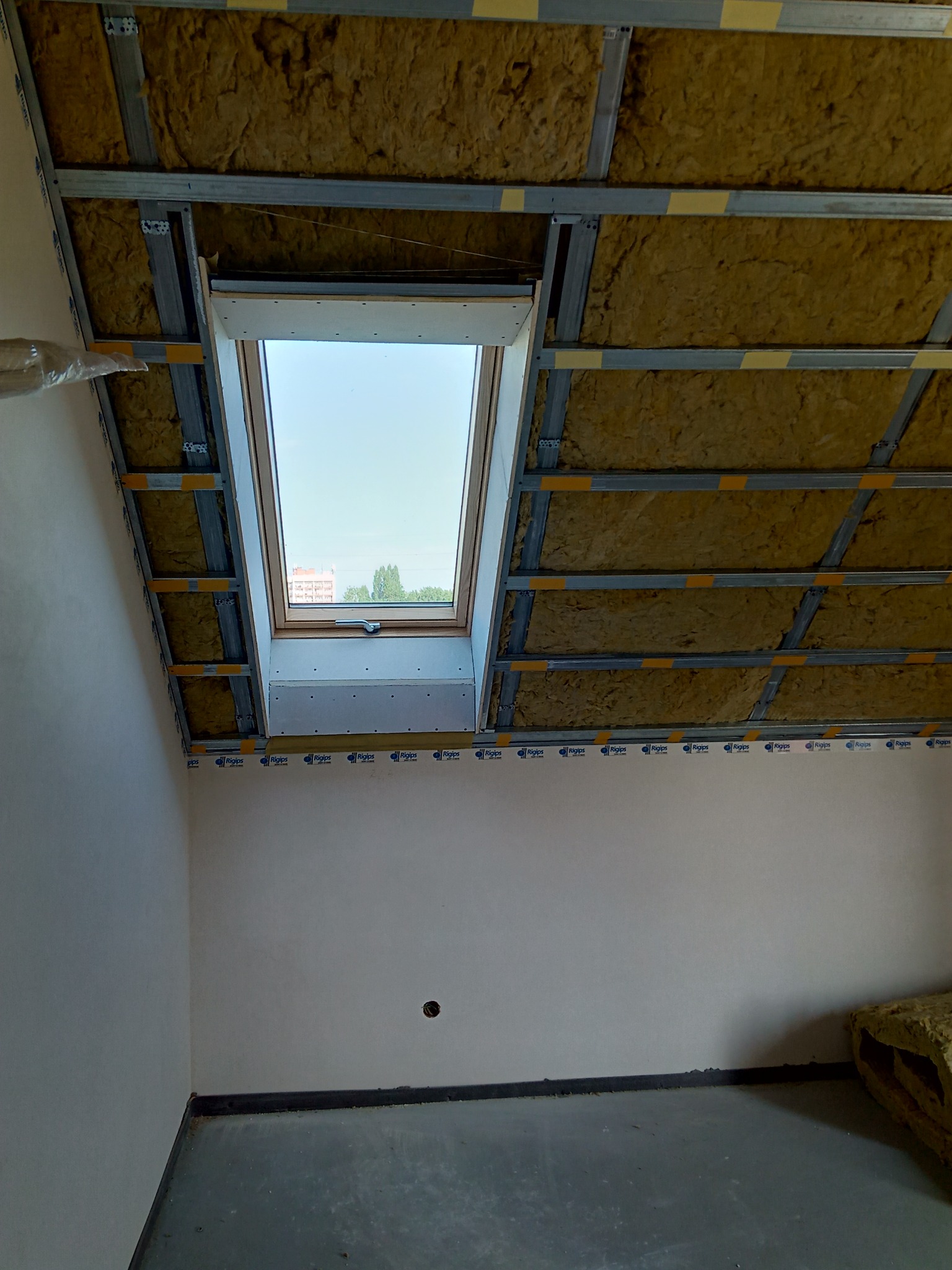 Montaż izolacji z wełny mineralnej w konstrukcji sufitu poddasza z oknem dachowym, widoczne metalowe profile i taśma uszczelniająca.