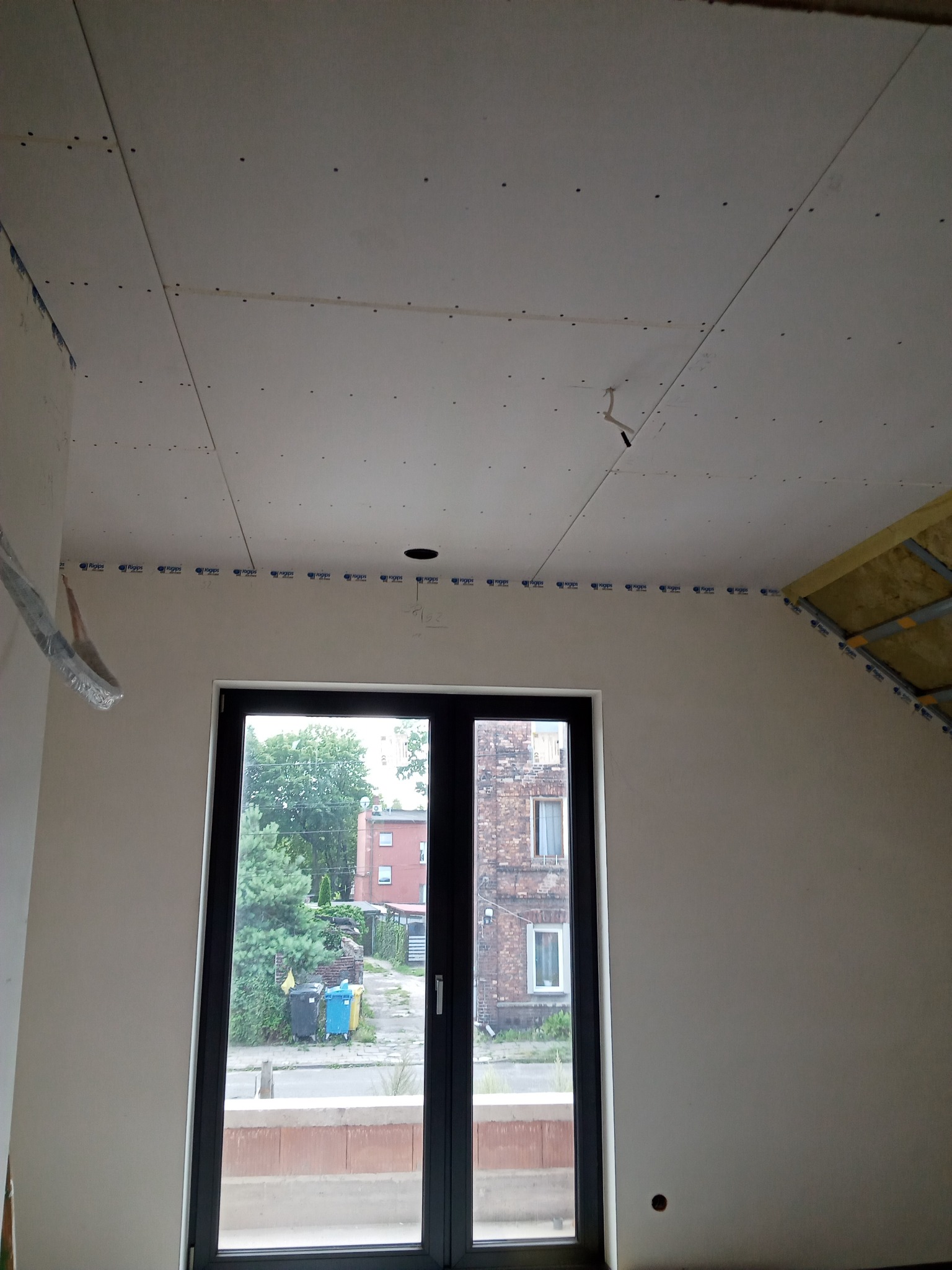 Wnętrze w trakcie remontu, widoczne zamontowane płyty na suficie, zabezpieczone taśmą, z widocznym otworem na oświetlenie, oraz fragment poddasza z izolacją, okno z widokiem na ulicę i budynki.