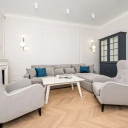 Jasne wnętrze salonu z parkietem w jodełkę, szarą sofą narożną z turkusowymi poduszkami, dwoma fotelami typu uszak, białym stolikiem kawowym, czarną witryną i dekoracyjnym kominkiem z wazonem...