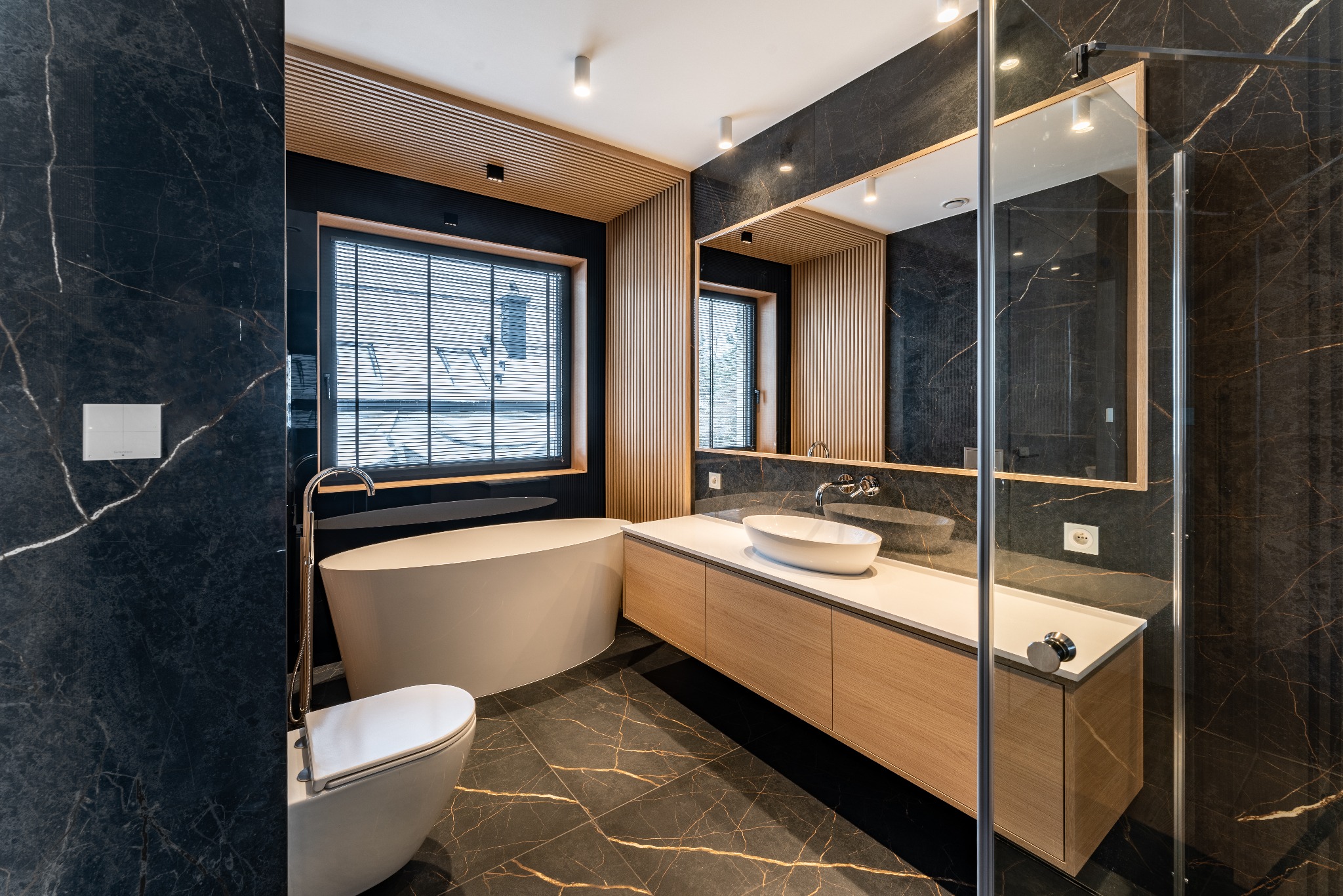 Nowoczesna łazienka z czarnymi marmurowymi ścianami i podłogą, jasnym drewnem na suficie i szafkach, owalną wanną i umywalką nablatową oraz przeszkloną kabiną prysznicową.