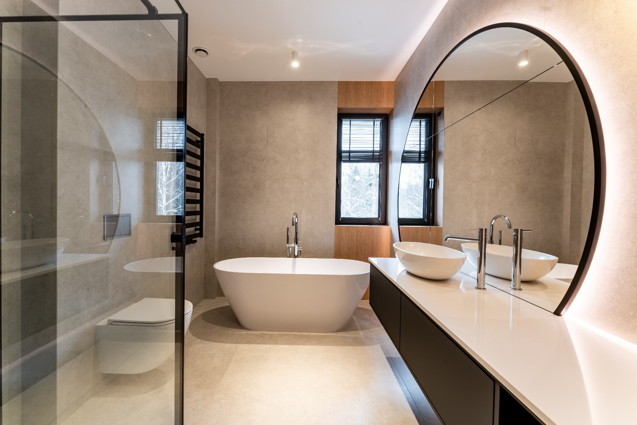 Nowoczesna łazienka z owalną wanną wolnostojącą, podwieszaną toaletą za szklaną ścianką, dwoma umywalkami nablatowymi i owalnym lustrem z podświetleniem LED.