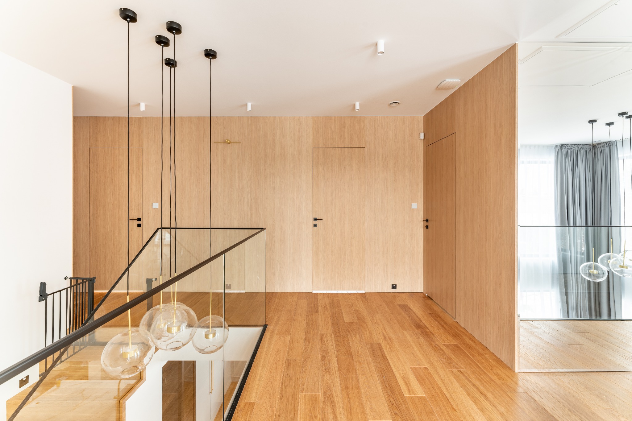 Nowoczesne wnętrze domu z drewnianą podłogą, ścianami wykończonymi fornirem i szklaną balustradą schodów, oświetlone wiszącymi lampami z kulistymi kloszami.