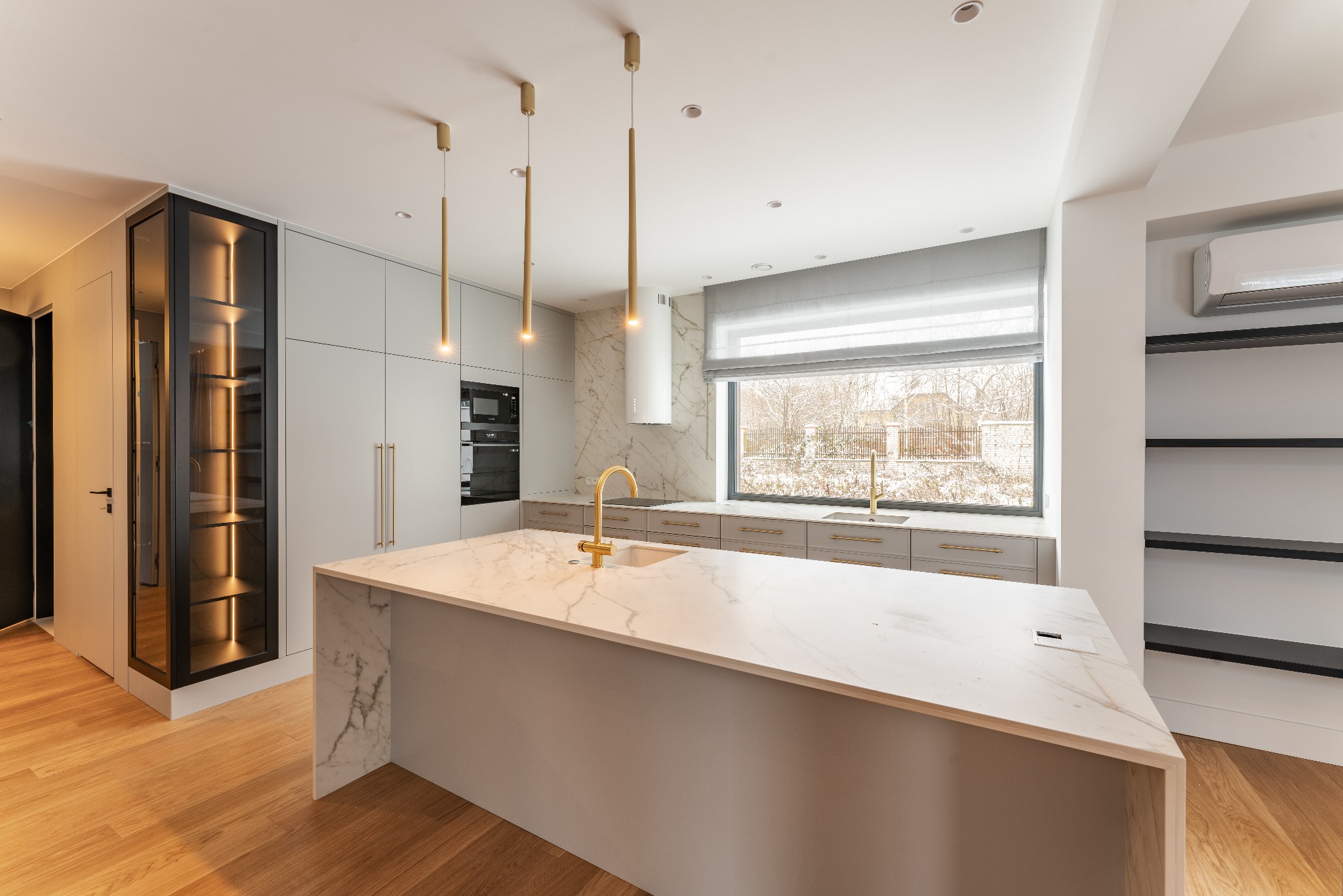 Nowoczesna kuchnia z marmurową wyspą, złotą armaturą i wiszącymi lampami, oświetlona naturalnym światłem wpadającym przez duże okno.