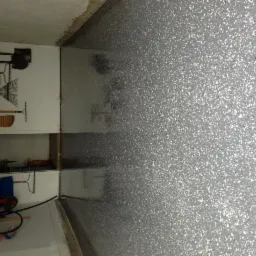 Szara, błyszcząca posadzka z drobnymi, kolorowymi elementami w garażu, widoczna ściana z narzędziami ogrodniczymi.