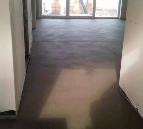 Gładka, szara powierzchnia wylewki w nowym budynku, widoczne okna z cegłami w tle, surowe wykończenie.