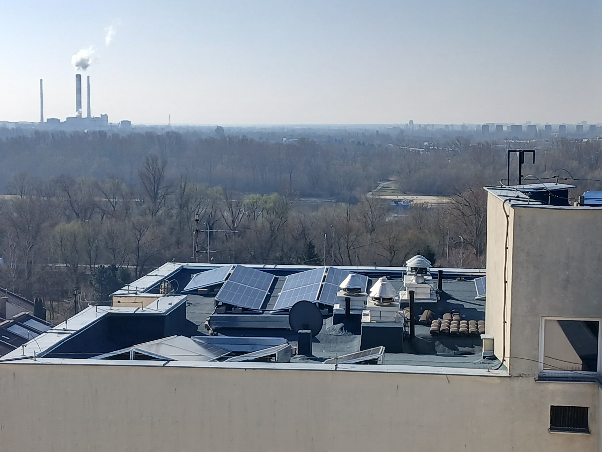 Instalacja paneli słonecznych na dachu budynku z widokiem na odległy krajobraz miejski i kominy elektrowni w tle.