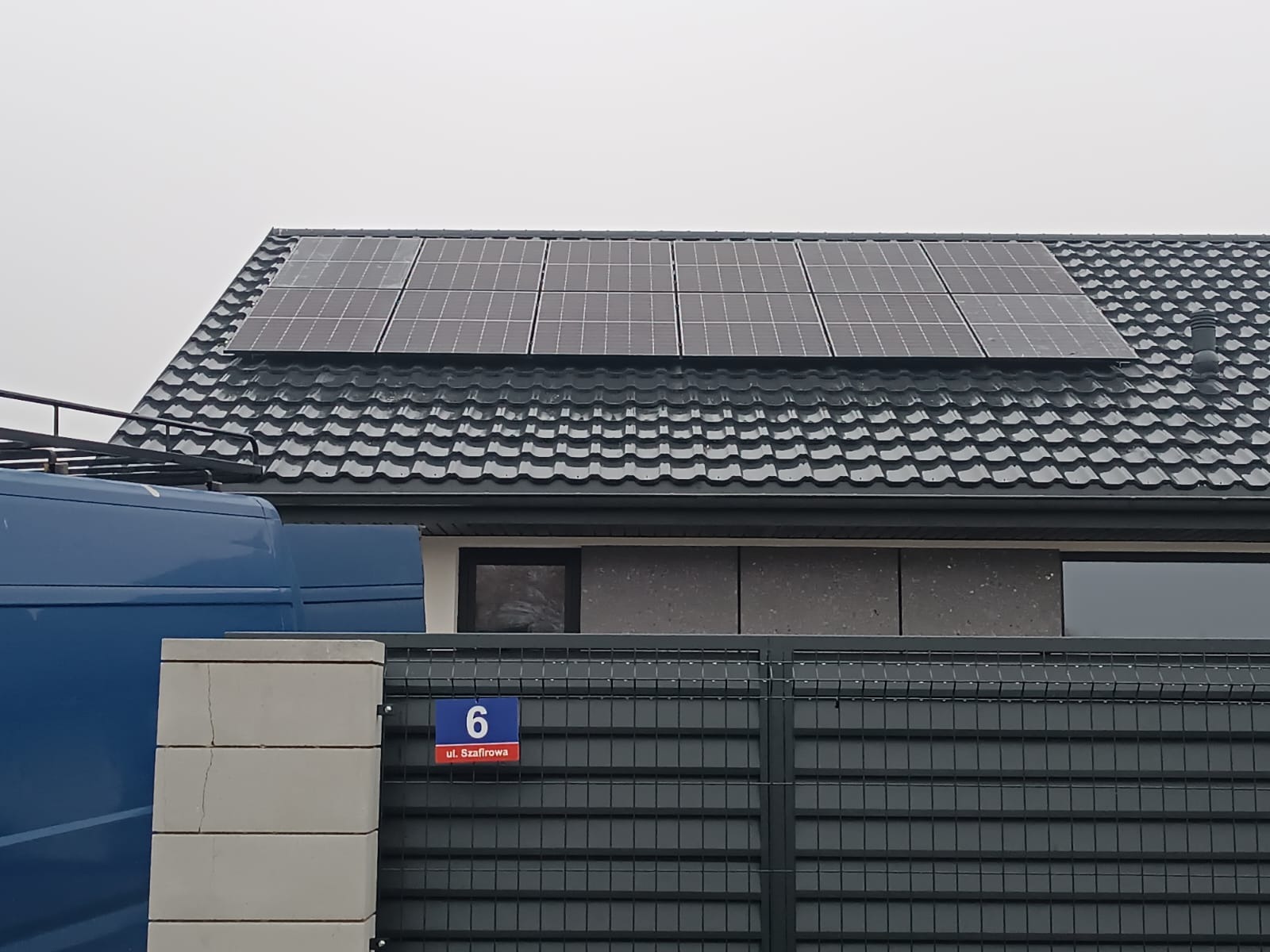 Panele słoneczne na ciemnym, metalowym dachu domu jednorodzinnego, widoczne zza szarego ogrodzenia z numerem 6 przy ulicy Szafirowej, z fragmentem niebieskiego samochodu dostawczego po lewej stronie.