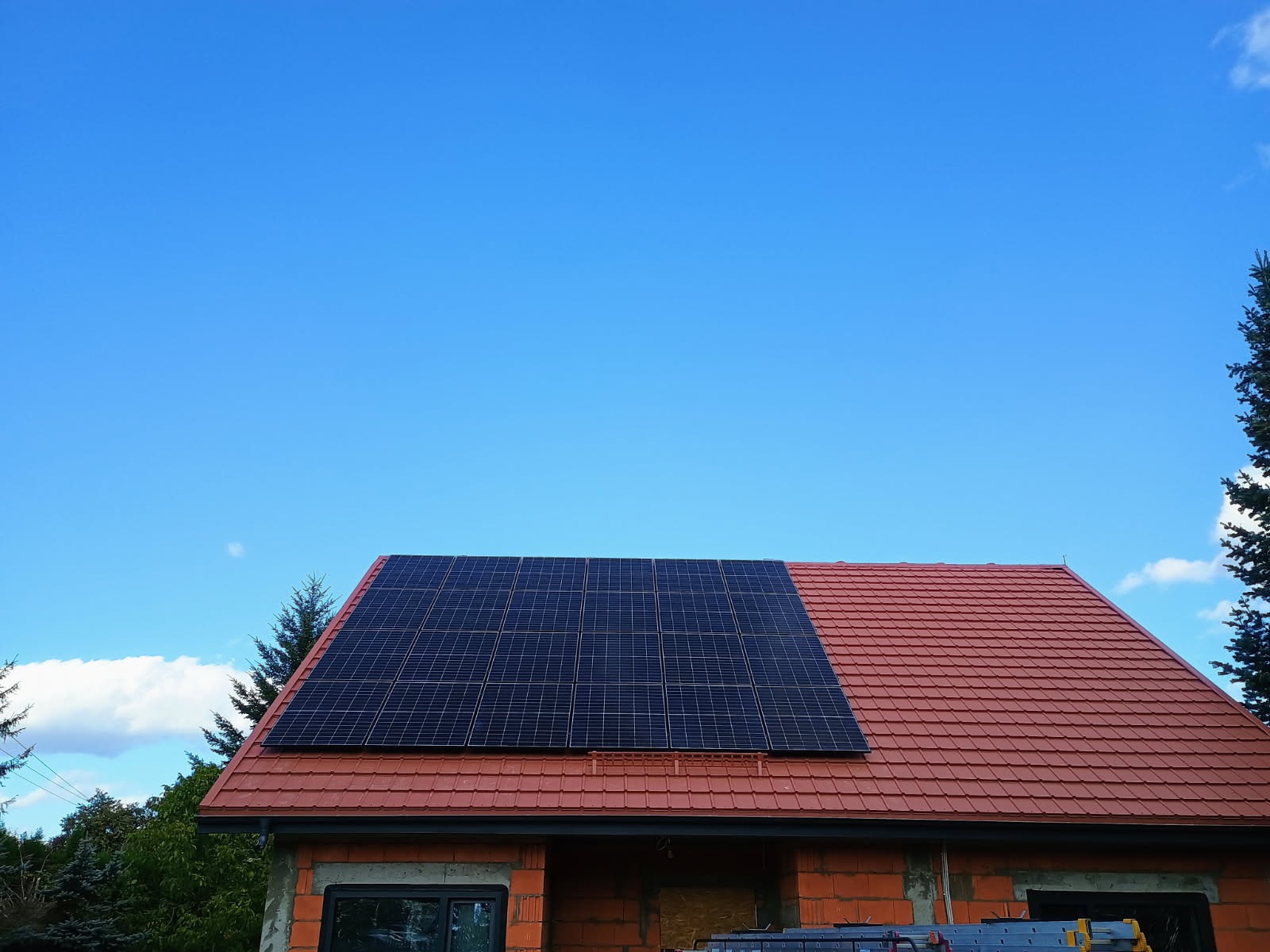 Częściowo zamontowane panele fotowoltaiczne na dachu domu z czerwoną dachówką w słoneczny dzień.