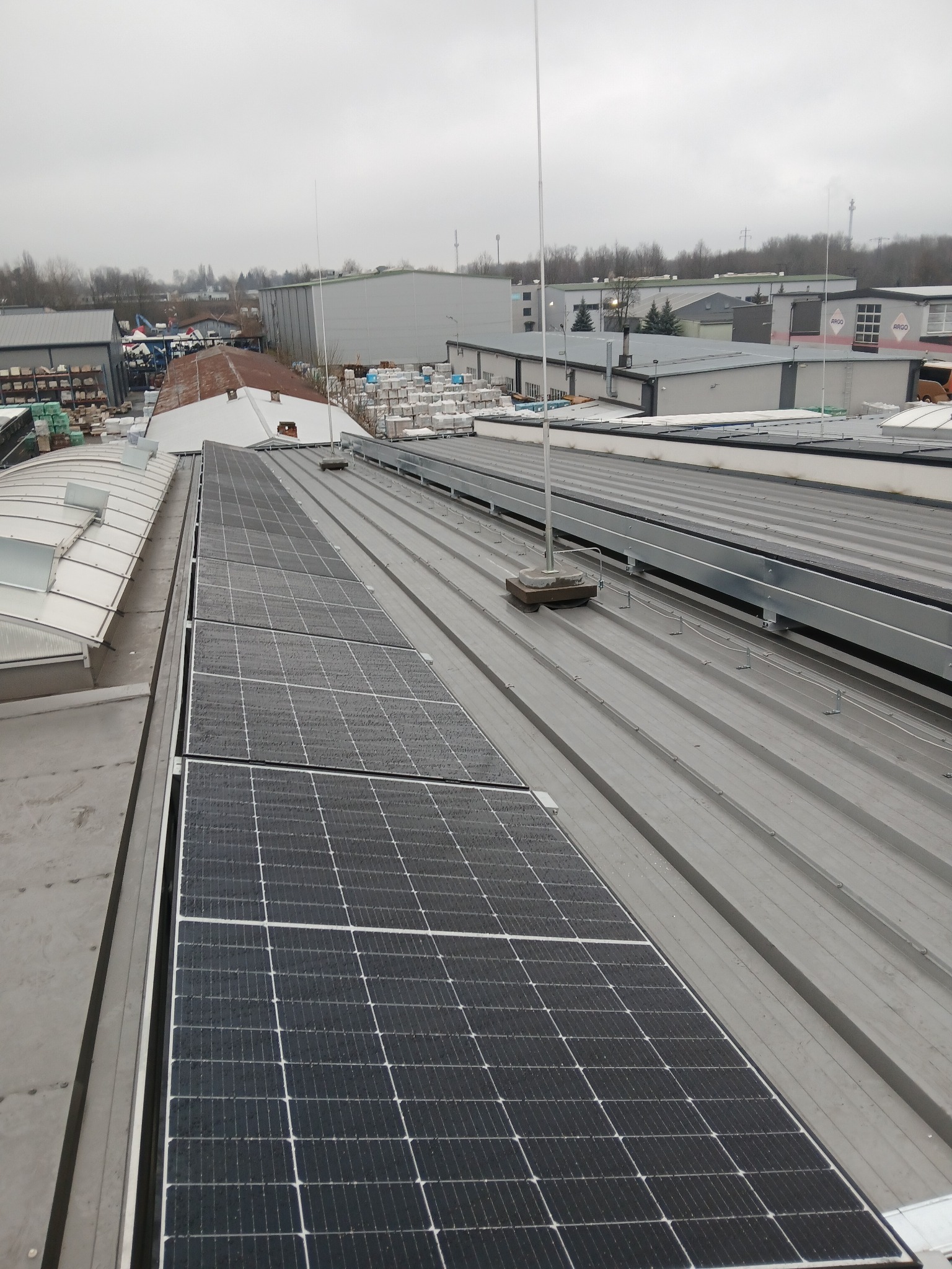 Rząd paneli słonecznych na metalowym dachu budynku przemysłowego w pochmurny dzień, widoczne elementy mocujące i odgromniki.