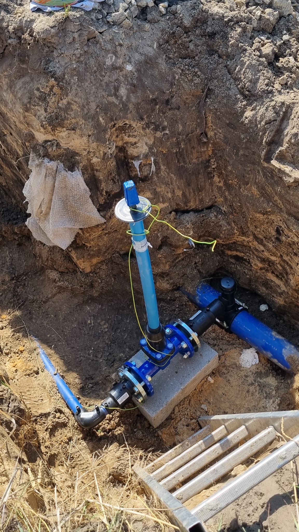 Niebieskie rury wodociągowe połączone z czarnym zaworem, zamontowane w wykopie na betonowym fundamencie, widoczne przewody elektryczne i drabina w tle.