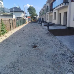 Przygotowanie podłoża pod chodnik z kostki brukowej przed szeregiem nowych domów, widoczne gruz i piasek, w tle maszyna budowlana.