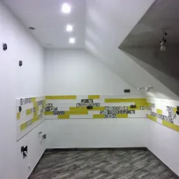 Wnętrze kuchni w trakcie wykańczania, z ułożonymi płytkami dekoracyjnymi w kolorze żółtym, białym i wzorzystym nad blatem, widoczne niedokończone instalacje elektryczne i hydrauliczne, oraz podłoga...