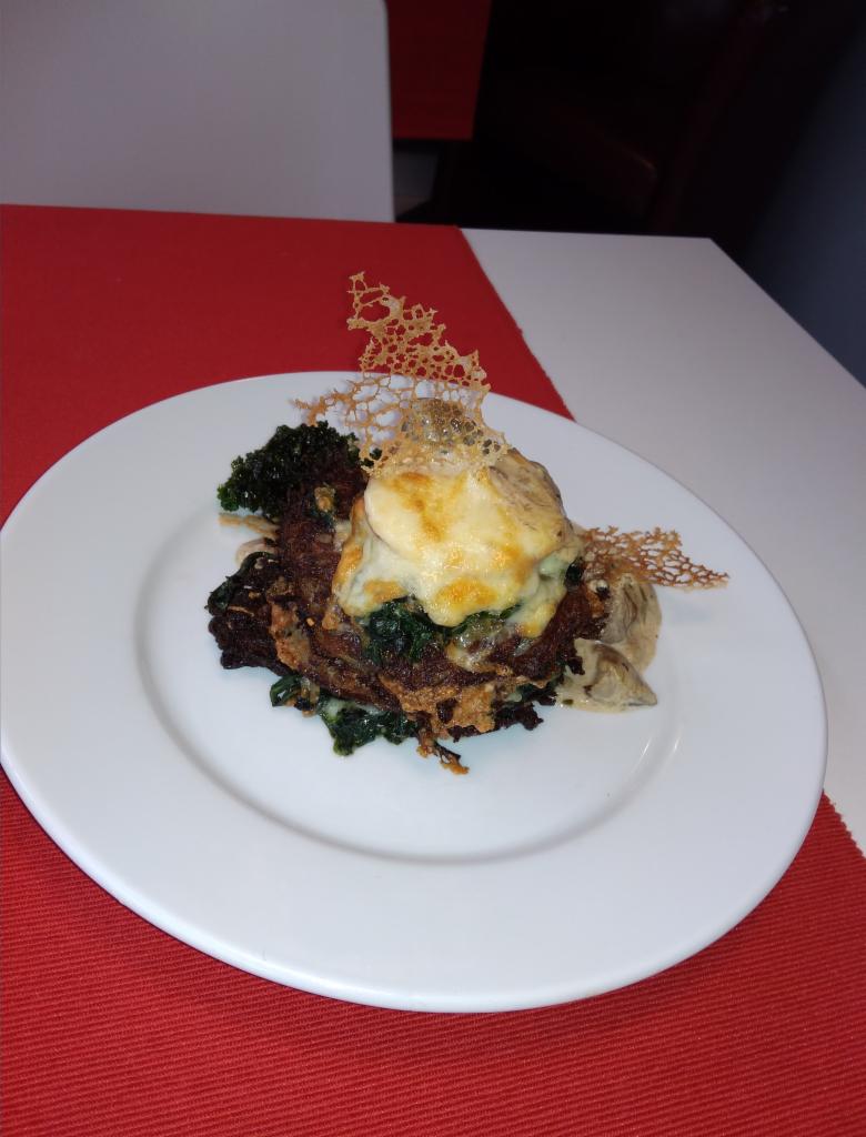 Elegancko podane danie na białym talerzu z dekoracją z karmelu, składające się z warstw placków ziemniaczanych, szpinaku i zapiekanego sera, na czerwono-białym tle.