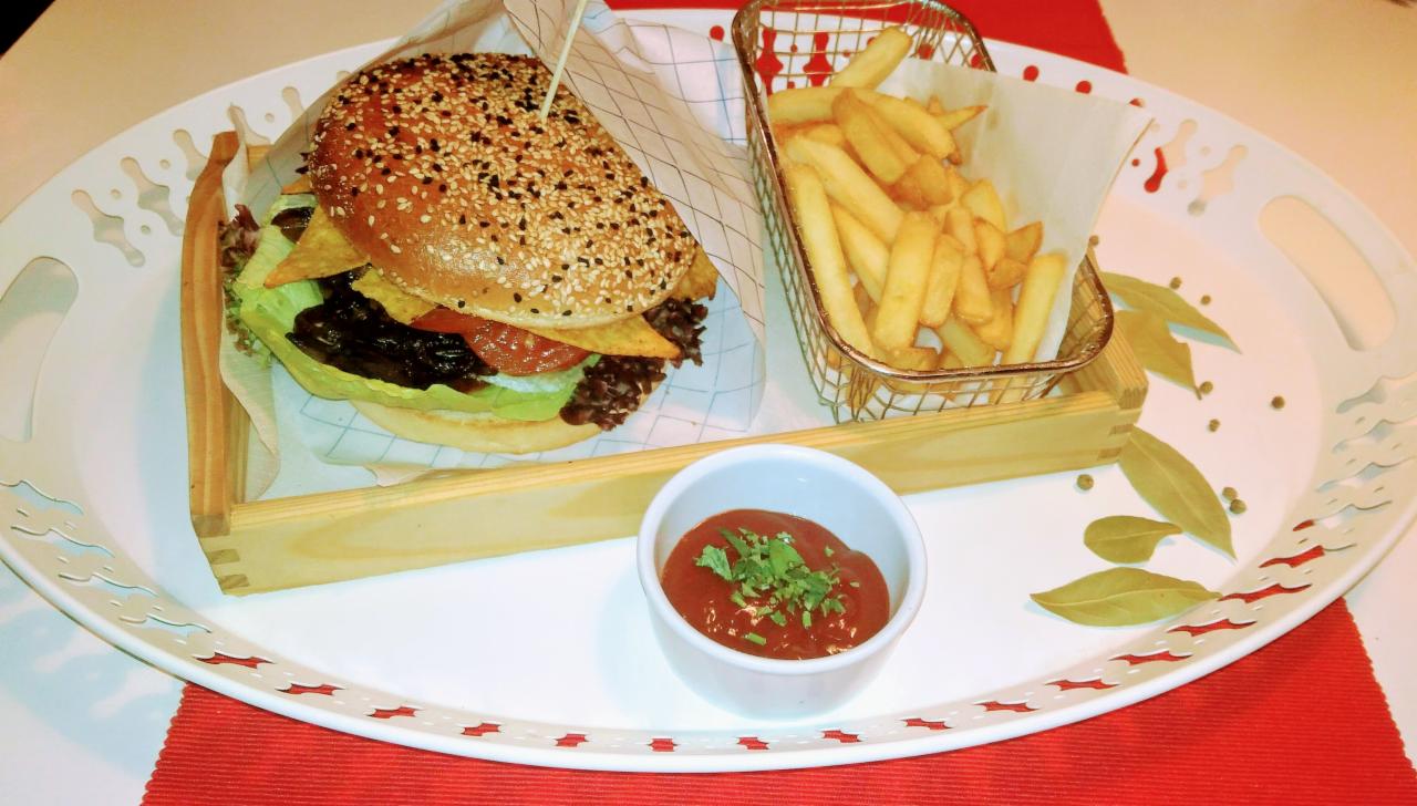 Burger z sezamową bułką, frytkami w metalowym koszyczku i sosem w białej miseczce, podane na ozdobnej tacy z drewnianymi elementami, na czerwonej podkładce.