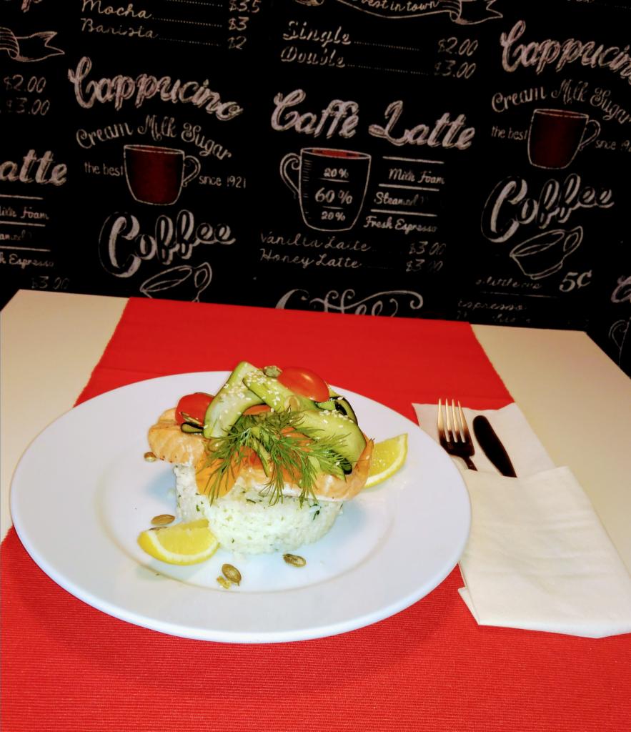 Elegancko podane danie: ryż z łososiem, ogórkiem, pomidorkami cherry, koperkiem i cytryną na białym talerzu, widelcem i nożem obok, na tle tablicy z menu kawiarni i czerwono-białego obrusu.