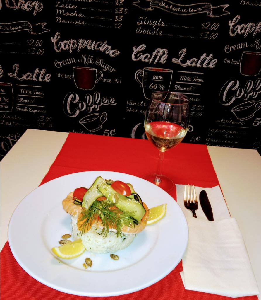 Elegancko podane danie z ryżu, łososia i warzyw na białym talerzu, z cytryną i kieliszkiem białego wina na stole z czerwoną serwetą, tło z motywami kawowymi.