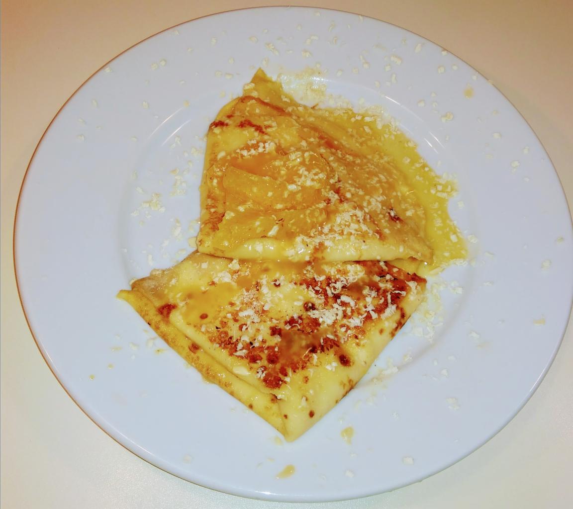 Dwa naleśniki złożone w trójkąty na białym talerzu, polane miodem i posypane wiórkami kokosowymi, widoczne z góry w naturalnym oświetleniu.