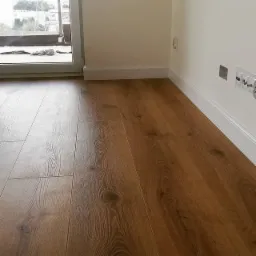 Ułożona podłoga z paneli w odcieniach brązu z widoczną strukturą drewna, białe listwy przypodłogowe przy jasnych ścianach, gniazdka elektryczne w tle.