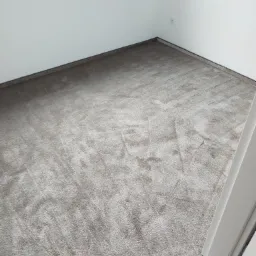 Jasnoszara wykładzina dywanowa ułożona w rogu pokoju z białymi ścianami i listwami przypodłogowymi, widoczny fragment ramy drzwiowej.