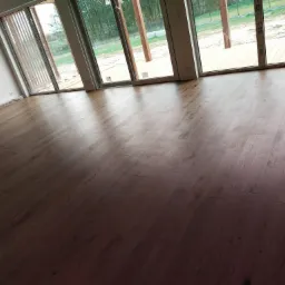 Ułożona, ciemna podłoga drewniana w nowym domu z dużymi oknami tarasowymi, widoczne listwy przypodłogowe z logo producenta i butelka wody w rogu pokoju.