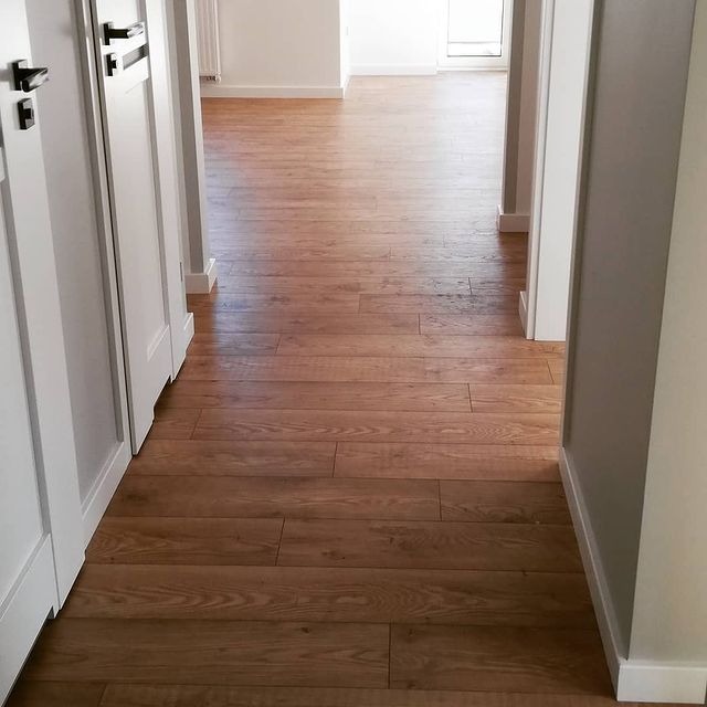 Korytarz z ułożoną jasnobrązową podłogą z desek, widoczne białe drzwi z czarnymi klamkami i szare ściany z białymi listwami przypodłogowymi.