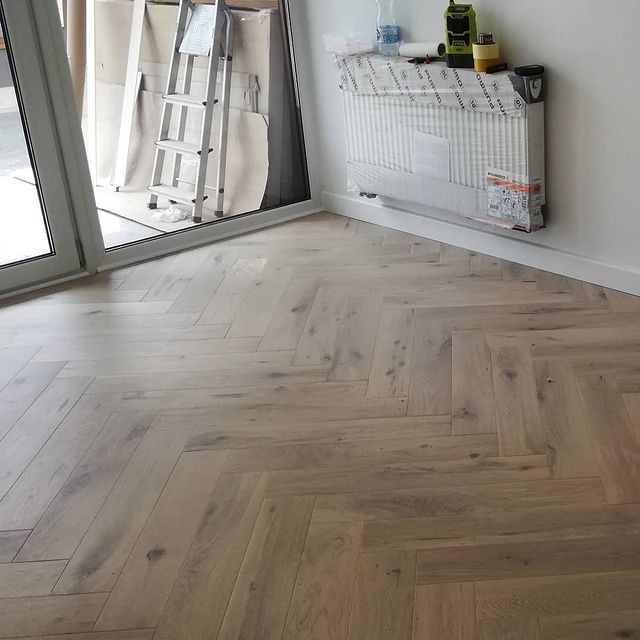 Ułożona jasna podłoga z desek w jodełkę francuską, widoczny fragment okna balkonowego, drabina oparta o ścianę i zabezpieczony folią grzejnik.