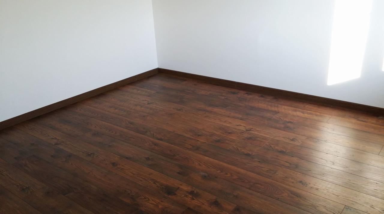 Ciemnobrązowa podłoga z paneli ułożona w rogu pomieszczenia z białymi ścianami i przypodłogowymi listwami w kolorze podłogi.