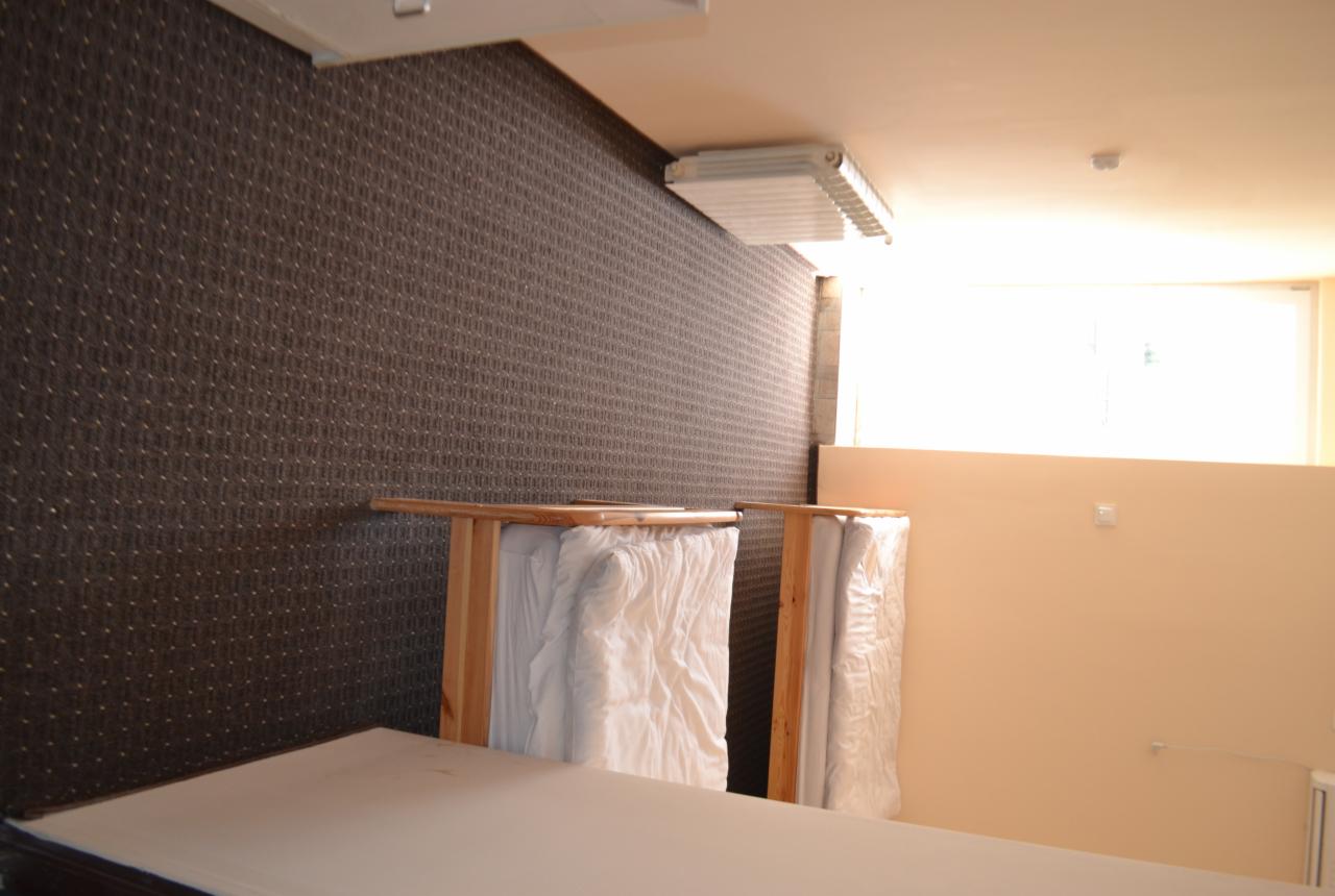 Przechowywane pionowo w drewnianych ramach, zabezpieczone folią, rolki wykładziny na tle ściany pokrytej wzorzystą tapetą.