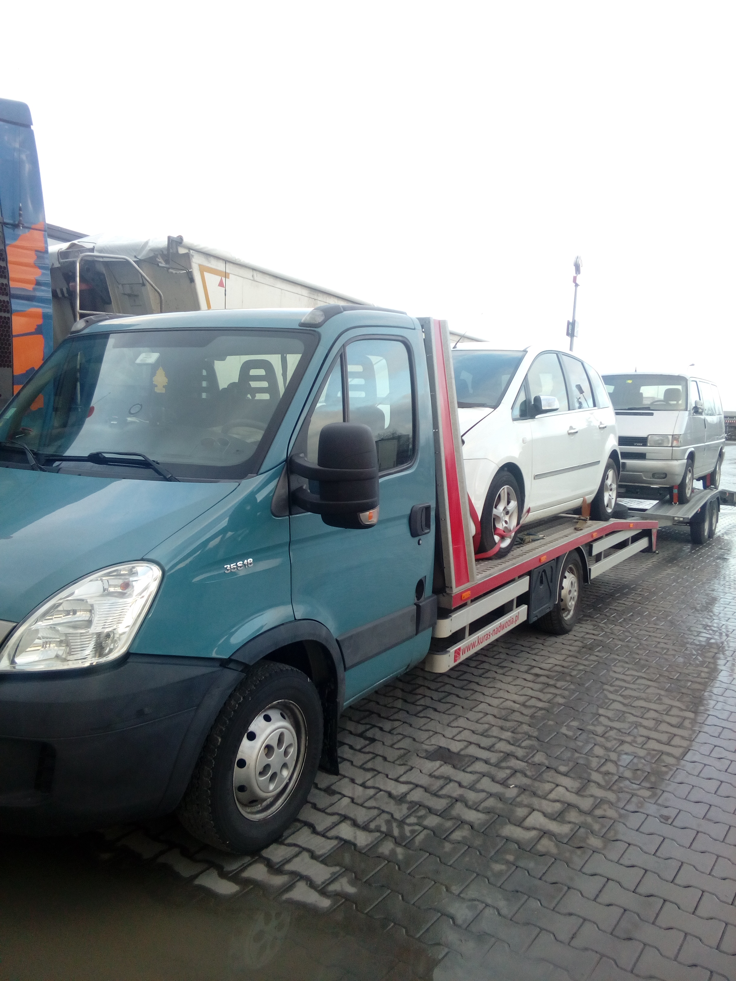 Turkusowy samochód z platformą transportową przewozi dwa samochody osobowe na mokrej kostce brukowej.