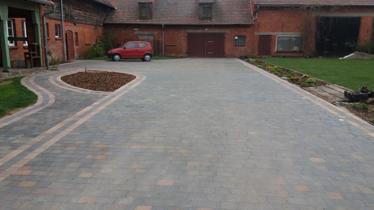 Szeroki podjazd z kostki brukowej w odcieniach szarości i beżu, obrzeża wykończone jaśniejszą kostką, na tle ceglanego budynku gospodarczego i czerwonego samochodu.