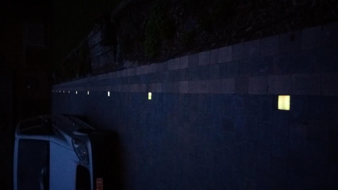 Oświetlona ściana z ciemnej kostki brukowej z wbudowanymi lampkami, nocna sceneria z zaparkowanym samochodem w tle.