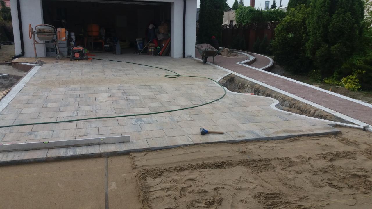 Fragment układanej nawierzchni z kostki brukowej w odcieniach szarości, widoczna poziomica i gumowy młotek, w tle mieszalniki do betonu i częściowo ułożony chodnik z czerwonej kostki.