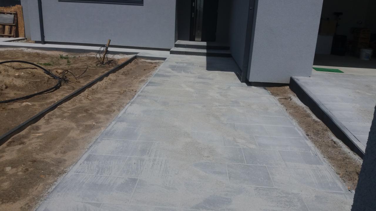 Świeżo ułożona, szara kostka brukowa prowadząca do wejścia domu, z widocznymi obrzeżami i ziemią po bokach, jeszcze przed ostatecznym wykończeniem.