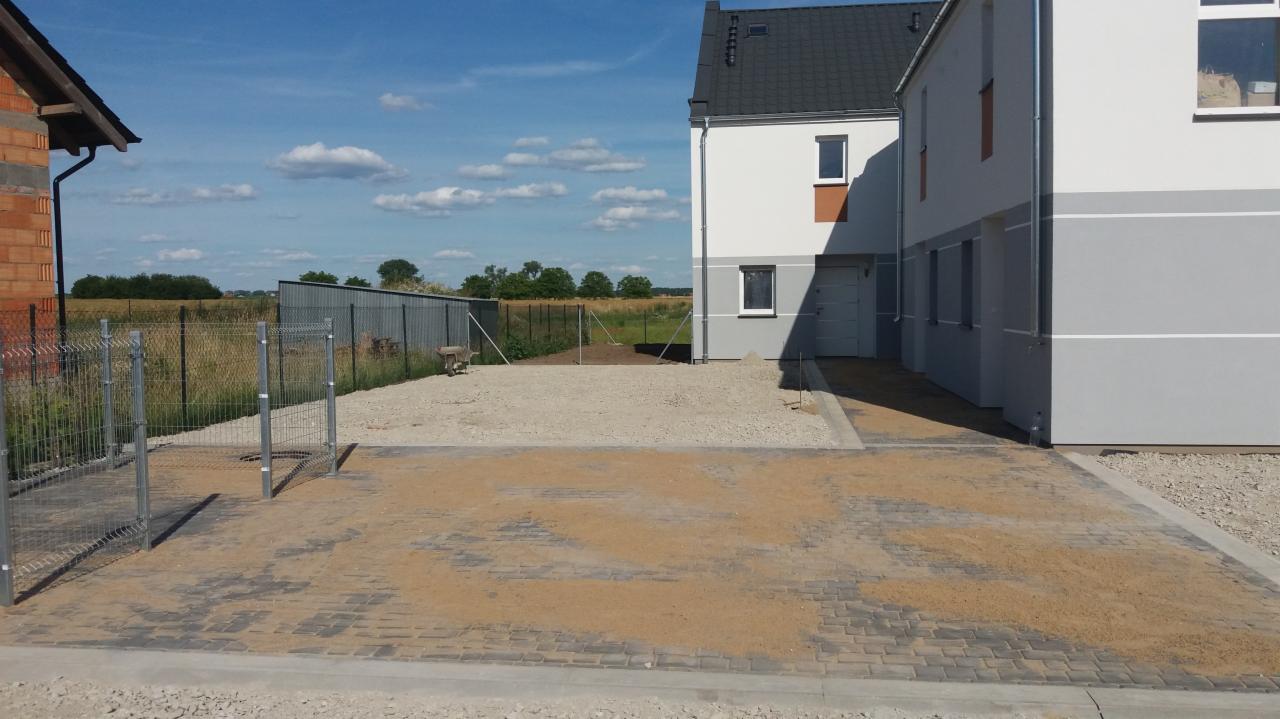 Nowo ułożona, szara kostka brukowa, częściowo zasypana piaskiem, na podjeździe przed nowoczesnym domem w biało-szarej kolorystyce, widoczny fragment ogrodzenia i przygotowany teren obok.