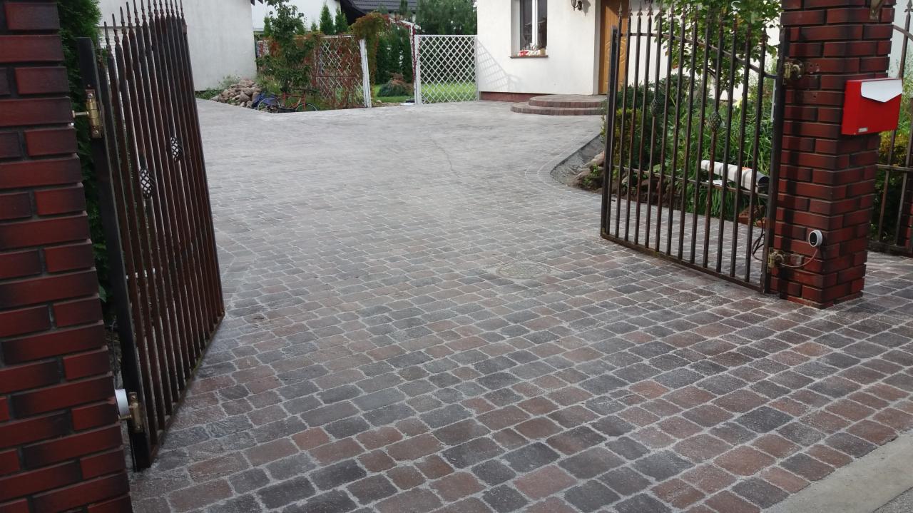 Ułożona kostka brukowa w odcieniach szarości i brązu tworząca podjazd, widoczna brama wjazdowa z metalowymi prętami i murowanymi słupkami z czerwoną skrzynką na listy.