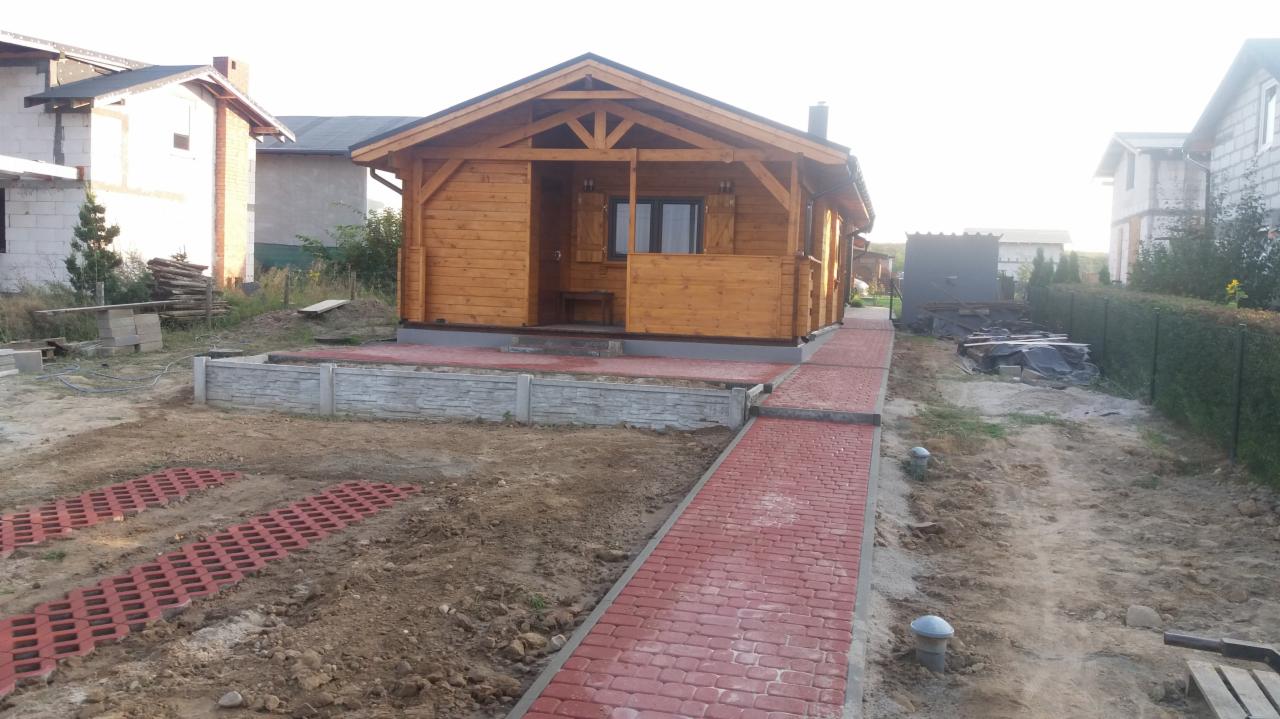Nowo wybudowany, drewniany domek letniskowy z czerwonym chodnikiem z kostki, otoczony przygotowanym terenem pod trawnik i widocznymi elementami betonowymi.