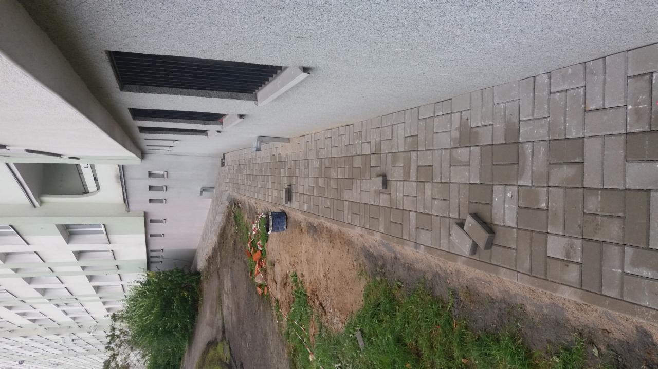 Ułożona kostka brukowa wzdłuż budynku, widoczne pozostałości prac ziemnych i wiadro, dwie kostki leżące na boku.