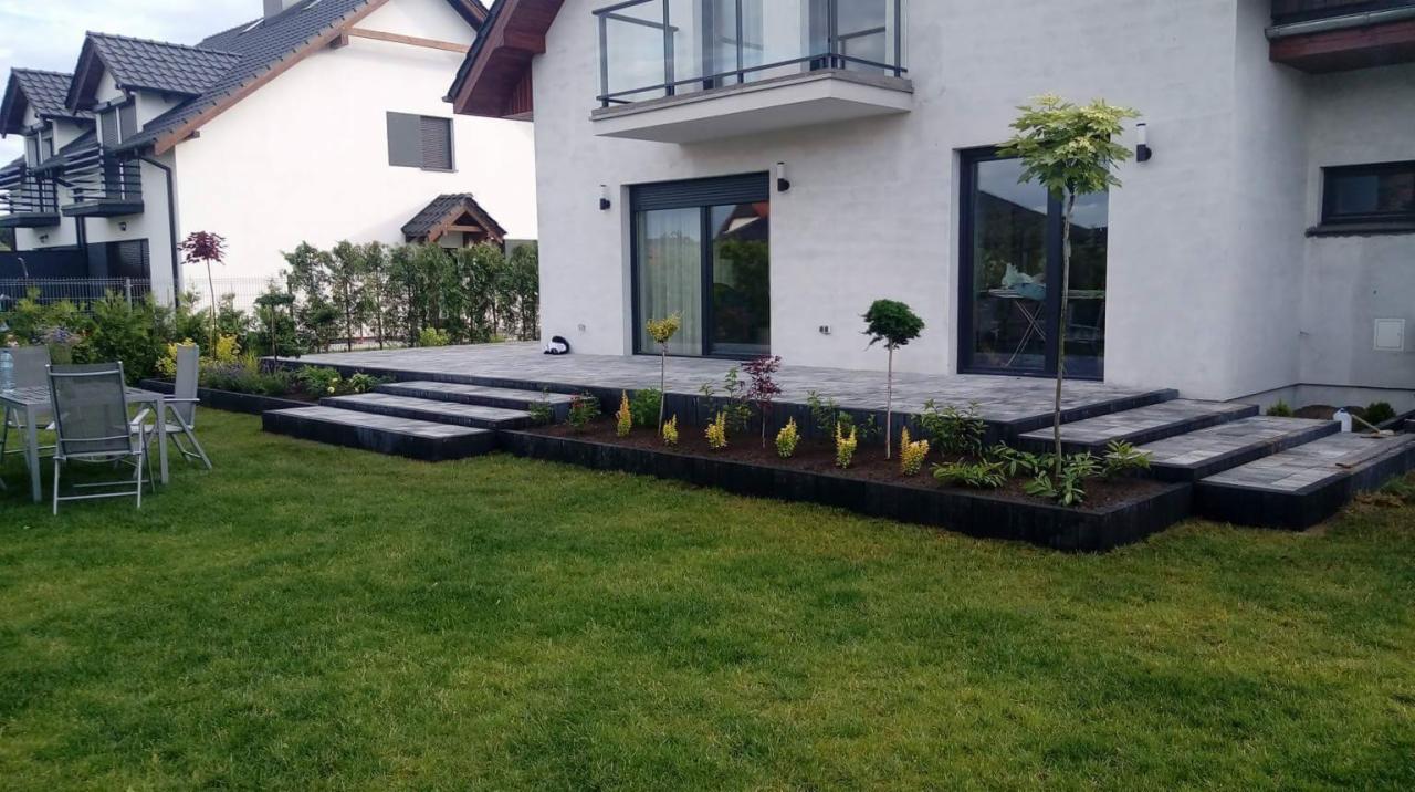 Ułożona szara kostka brukowa tworząca taras i schody przed nowoczesnym domem z ogrodem, widoczne nasadzenia roślin wzdłuż obrzeża, zielony trawnik.