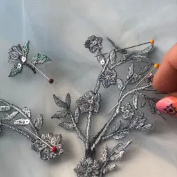 Delikatna, srebrna aplikacja z motywem kwiatowym i cekinami, przypięta szpilkami do jasnoniebieskiego tiulu, z widoczną dłonią z pomalowanymi paznokciami.