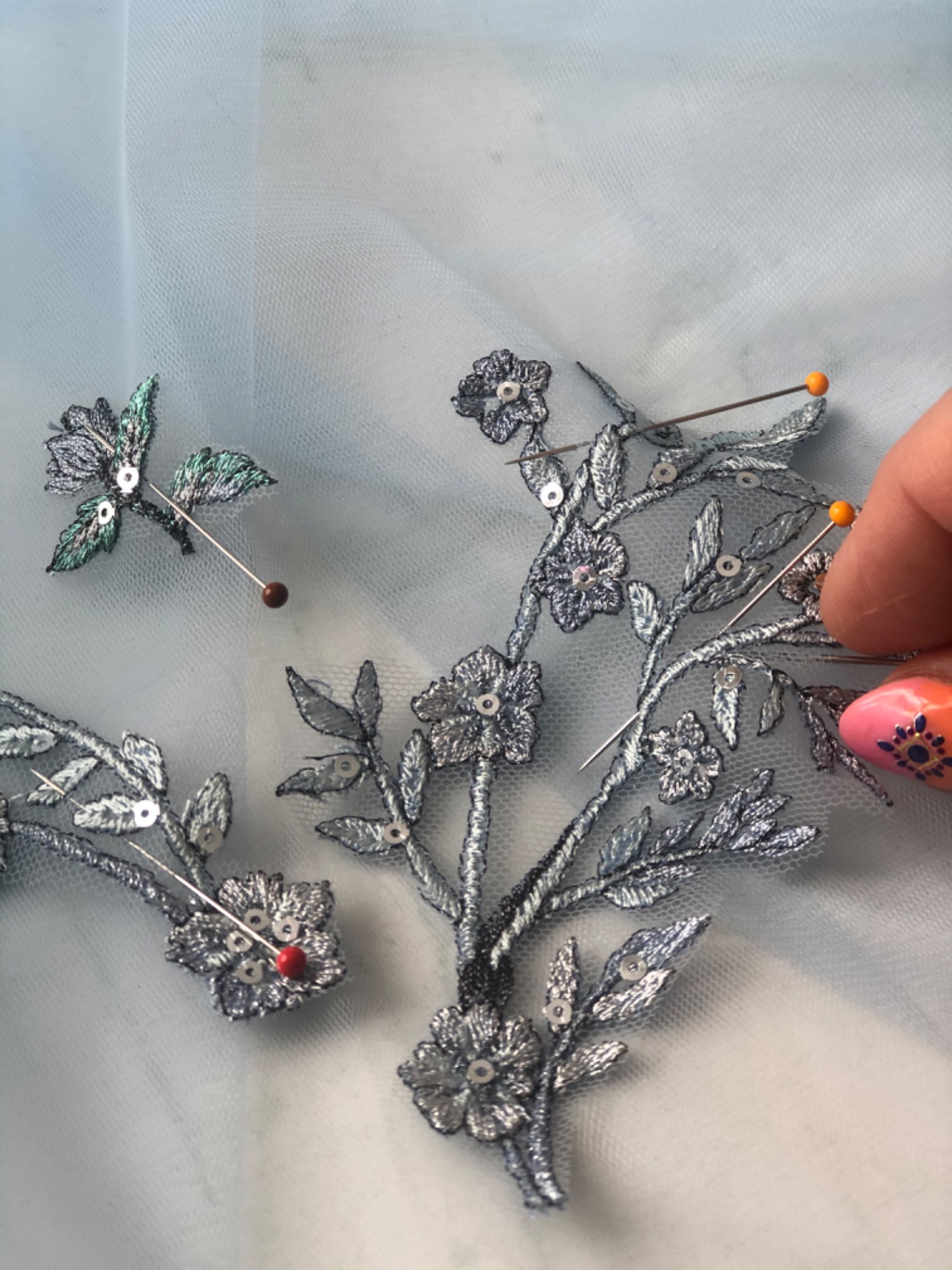 Delikatna, srebrna aplikacja z motywem kwiatowym i cekinami, przypięta szpilkami do jasnoniebieskiego tiulu, z widoczną dłonią z pomalowanymi paznokciami.